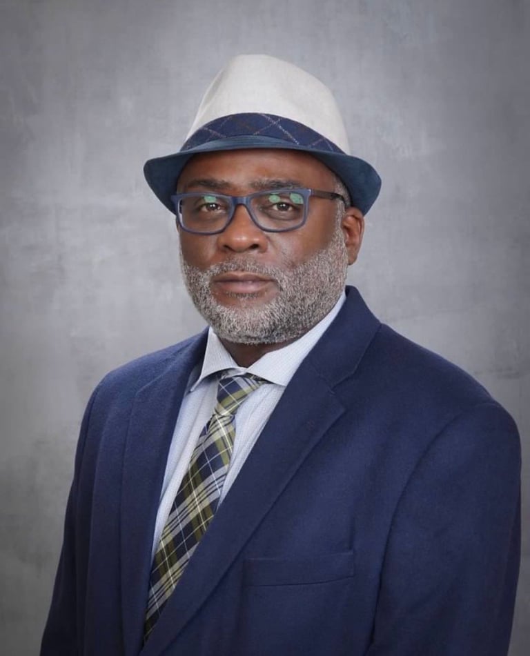 Professional headshot of James Theodore Robinson Jr. wearing a navy blazer, plaid tie, glasses, and a navy and white fedora hat